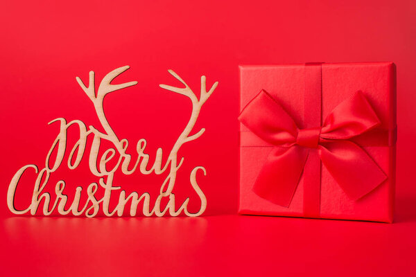 Christmas is coming concept. Close up photo of red giftbox with red ribbon and merry chrismas wooden curved decoration isolated on red background