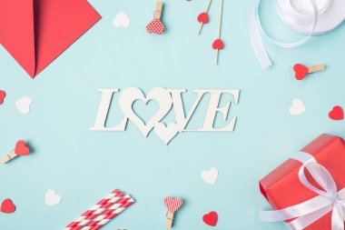 Yukarıdaki fotoğrafta zarf raptiyeleri küçük kalpler ve açık mavi arkaplan üzerine paketlenmiş hediye kutusu var.