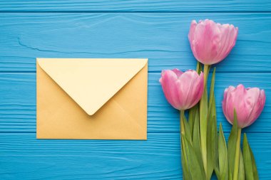 En üstte yeşil saplı güzel çiçeklerin ve parlak mavi masadaki altın renkli kağıt zarfların olduğu düz bir resim var.