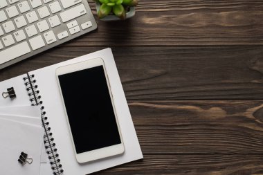 Üst tepedeki görüntüde bilgisayar klavyesinin spiral klasör not defterindeki bitkisel açık not defteriyle fotoğrafı ve cep telefonunda karanlık ahşap zemin üzerinde boş kopya alanı bulunan boş bir cep telefonu.