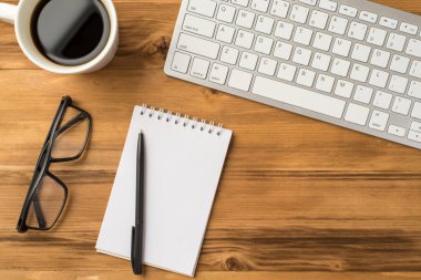 Tepede bilgisayar klavyesinin, sabah içecek kadehinin, gözlüklerin ve ahşap zemin üzerindeki sarmal not defterinin yakın görüntüsü var.