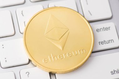 Üst tepede, arka planda beyaz dizüstü bilgisayarın klavyesinde duran parlak altın etherium 'un yakın çekim fotoğrafı var.