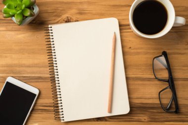 Yukarının üstünde boş bir defterin, açık bir kupanın, kahverengi ahşap zemin üzerinde yeni içecekten yapılmış cep telefonu ve gözlüklerin resmi var.