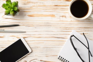 Yukarda, gözlüklü fincanın yakın plan fotoğrafı. Dijital telefon çiçeği saksısı ve telif defteri. Boş alan ile izole edilmiş beyaz ahşap zemin.