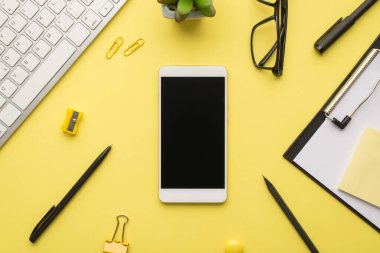 Telefon, klavye, kağıt ataç kalem, bileyici ve gözlüklerin üstünde parlak sarı arkaplan görüntüsü.