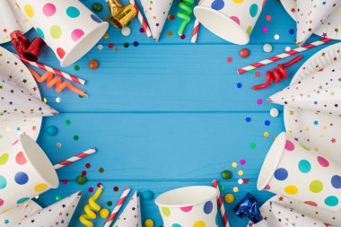 Doğum günü partisi kompozisyonu, spiral mumlar, kamışlı şapkalar, konfeti polka noktaları, kağıt bardaklar ve kopyalanmış mavi ahşap masa arka planında tabaklar.