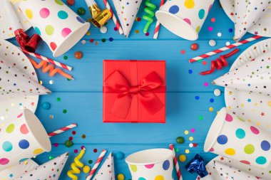 Kırmızı hediye kutusunun en üstteki fotoğrafı, doğum günü kompozisyonu ortasında kurdele ve izole edilmiş mavi ahşap masa arka planında parti aksesuarları.