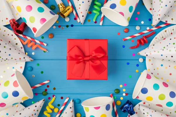 Kırmızı hediye kutusunun en üstteki fotoğrafı, doğum günü kompozisyonu ortasında kurdele ve izole edilmiş mavi ahşap masa arka planında parti aksesuarları.