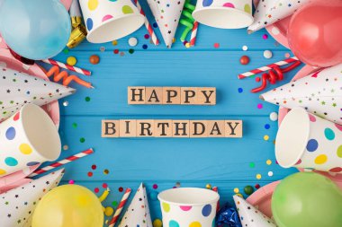 Doğum günü kompozisyonu ahşap küplerin en üstteki fotoğrafı. Mutlu yıllar etiketli. Orta puantiyeli kağıt bardaklar ve pembe plakalı parti aksesuarları.