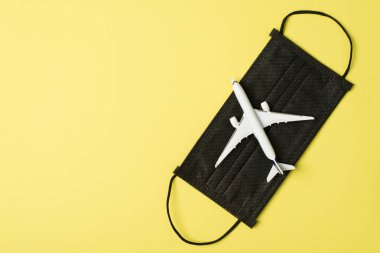 Siyah tıbbi yüz maskesi ve izole edilmiş pastel sarı arkaplan üzerindeki uçak modelinin fotosu.