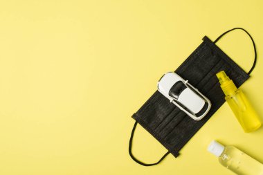 Siyah tıbbi yüz maskesi, sarı ve şeffaf dezenfektan şişeler, izole edilmiş pastel sarı arka plan, kopyalama alanı olan araba modelinin üst görüntüsü.