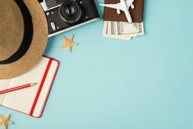 Güneş şapkalı kamera kırmızı kalem planlayıcısı ve pasaport kapağındaki uçak modelinin fotosu. Kopyalama alanı olan izole edilmiş pastel mavi arka planda dolar var.