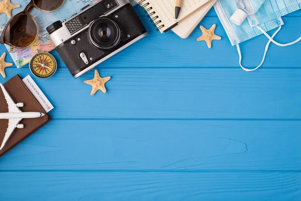 Above photo of camera plane compass map starfish notebook pen sanitizer sunglasses mask and passport with covid-19 test inside isolated on the blue wooden background with empty space