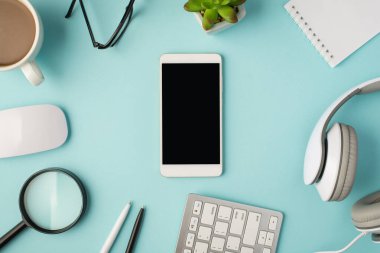 Orta fincandaki akıllı telefonun üst görünüm fotoğrafı faredeki kalemler büyüteç kulaklıklar not defterleri ve izole edilmiş pastel mavi arka plan üzerindeki gözlükler