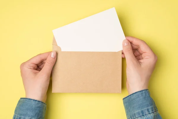 一人目の女性の手の写真は コピースペースと隔離された黄色の背景に白い紙のカードとオープンクラフト紙の封筒を保持 — ストック写真