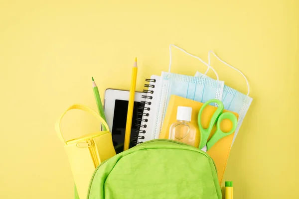 Above photo of backpack notepad pen pencil tablet pencil-case mask and sanitizer isolated on the yellow background