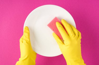 Sarı lastik eldivenli, beyaz, temiz plaka ve pembe arka planda pembe temizleme yastığı tutan ellerin en üstteki fotoğrafı.