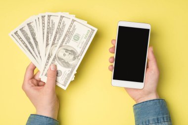 Para hayranı kadınların el resimleri. 100 dolarlık banknotlar ve cep telefonları. Kopyalanmış sarı arka planda.