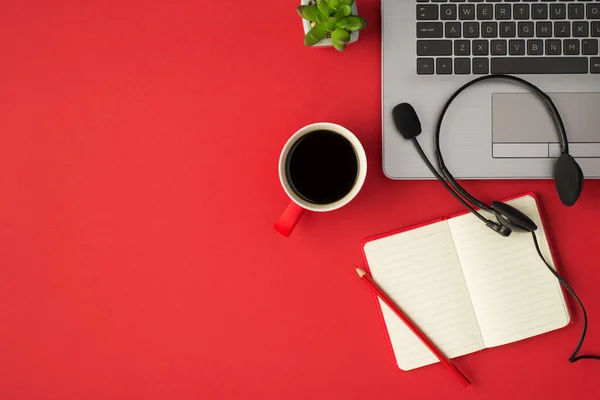 Top view photo of laptop black headset with microphone plant red cup of coffee and pencil on red planner on isolated red background with blank space