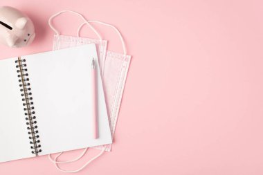 Üst görüntüde iki pembe tıbbi maske açık bir defter kalem ve izole edilmiş pastel pembe arka planda domuz kumbarası.