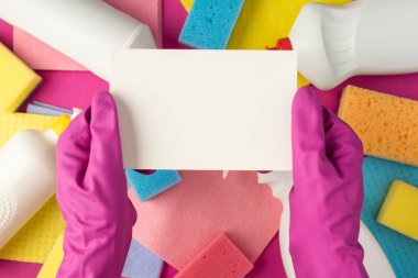 Üst görüntüde pembe lastik eldivenli eller beyaz kağıt kutu deterjan şişeleri tutarken çok renkli peçeteler kopyalanmış pembe arka planda temizleniyor.