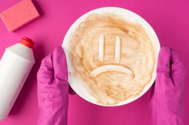 Üst görüntüde pembe lastik eldivenli ellerin kirli tabağı tutarken çekilmiş pembe yüz süngeri ve etiketsiz deterjan köpük şişesi izole edilmiş pembe arka planda boş alan