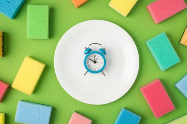 Ortadaki beyaz tabakta mavi alarm saatinin ve izole edilmiş yeşil arkaplanda çok renkli temizlik yastıklarının en üstteki görüntüsü