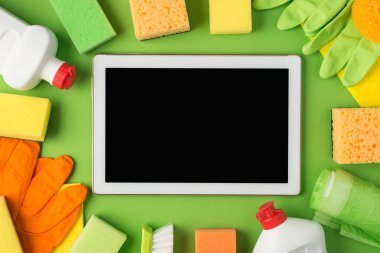 Ortadaki deterjan şişelerindeki tablet bilgisayarın en üst görüntüsü. Plastik eldivenler, süngerler, süngerler ve izole edilmiş yeşil arka planda çöp torbaları.