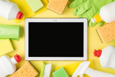 Ortadaki deterjan spreyindeki tablet bilgisayarın üst görüntüsü ve jel şişeleri sarı ve yeşil süngerler çöp poşetleri, çöp poşetleri ve izole edilmiş sarı arka planda plastik eldivenler.