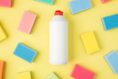 Etiketsiz beyaz deterjan şişesi ve izole edilmiş sarı zemin üzerindeki çok renkli süngerlerin üst görünüm fotoğrafı.