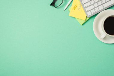 İşyeri klavyesinin en üstteki fotoğrafı yapışkan not kağıdı ataçları kalem bardakları ve içecekler tenha turkuaz arka planda boş arkaplanda.