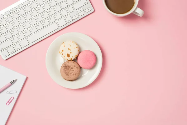 Above photo of keyboard cup of coffee notebook with pen paper clips and plate of various macaroons isolated on the pink backdrop with copyspace