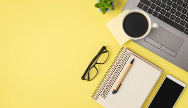 En üstteki fotoğrafta, dizüstü bilgisayar gözlükleri üzerinde sarı etiket kağıdı, çiçek saksısı akıllı telefon ve iki spiral defter, izole edilmiş pastel sarı arka plan ve kopyalanmış