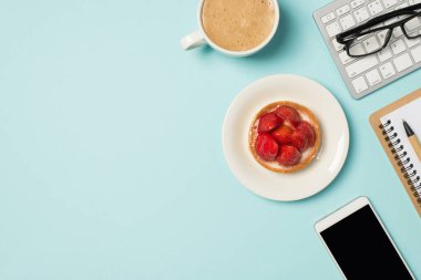 Klavye kalem düzenleyici tabağındaki gözlüklerin üst görüntüsü ahudududulu cep telefonu ve izole edilmiş pastel mavi arka planda bir fincan köpüklü latte.