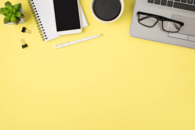 Dizüstü bilgisayarın, içki kadehinin, kalem klasörünün ve akıllı telefonun, izole edilmiş pastel sarı arka planda, boş alanlı, sarmal not defterinin en üst görüntüsü.
