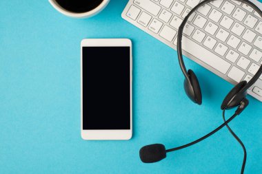 Üzerinde mikrofon olan siyah bir kulaklık, beyaz bir kahve fincanı ve cep telefonu, izole edilmiş açık mavi bir arkaplan ve boş bir alan.