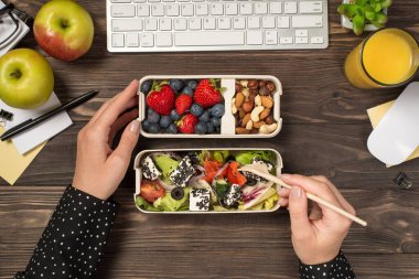 Ellerin sağlıklı yemek yediği, salata yediği, iki beslenme çantasından fındık yediği, elma bardağının, meyve suyu, kırtasiye malzemeleri, klavye ve farenin izole edilmiş, ahşap masa arkasında çekildiği ilk kişi.
