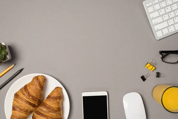 Top view photo of workspace keyboard mouse glasses binder clips cup of orange juice smartphone plate with croissants pencils and plant on isolated grey background with empty space