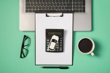 Dizüstü bilgisayarın, klavyenin, kahve fincanının, hesap makinesinin ve izole edilmiş turkuaz arka plandaki siyah klasörün en üstteki fotoğrafı.