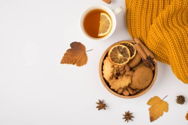 Sarı örülmüş eşarp sarı sonbahar yaprakları çay fincanı ve ahşap kase kurabiyeler badem fıstığı kurutulmuş limon dilimleri ve izole edilmiş beyaz arka planda tarçın çubukları.