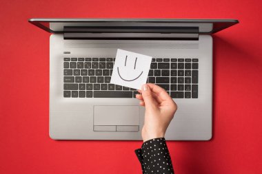 Üstten bakıldığında, kırmızı arka planda açık gri dizüstü bilgisayarın üzerine çizilmiş gülen yüzlü beyaz etiket kağıdı tutan ilk kişi.