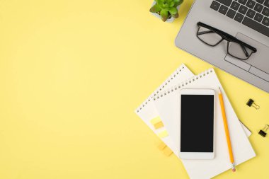 Dizüstü bilgisayardaki gözlüklerin en üstteki fotoğrafı. Kalem klasörü, cep telefonu ve iki sarmal not defteri. İzole edilmiş pastel sarı arka plan üzerinde sarı plastik yapıştırıcı yer imleri var.