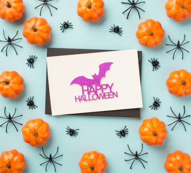 Cadılar Bayramı süslemelerinin en üstteki fotoğrafı siyah zarf simli yarasa ve yazıtlar beyaz kart balkabaklarında mutlu Cadılar Bayramı ve izole edilmiş pastel mavi arka planda örümcekler.