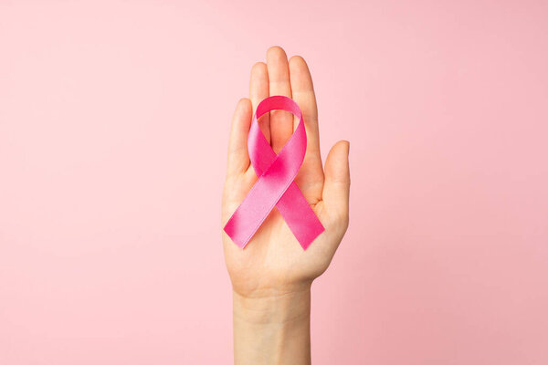 First person top view photo of hand holding pink ribbon in palm symbol of breast cancer awareness on isolated pastel pink background