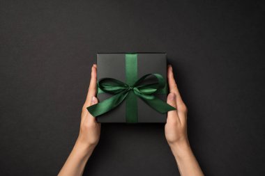 Siyah hediye kutusuyla yeşil saten kurdeleli siyah arka planda el ele tutuşan ilk kişinin fotoğrafı.