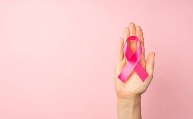 Avuç içi kanserinin sembolü olan pembe kurdeleli bir kadının, izole edilmiş pastel pembe arkaplanlı ve kopyalanmış pembe renkli bir el resmi.