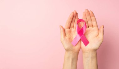 Üst düzey panoramik bir fotoğrafta, avuçlarında pembe kurdele tutan kadın elleri, izole edilmiş pastel pembe arka planda ve boş alanda meme kanseri farkındalığını simgeliyor.