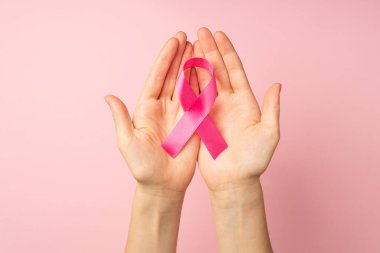 Göğüs kanserinin sembolü olan pembe kurdeleli ellerin izole edilmiş pastel pembe arka planda görüldüğü ilk kişi.