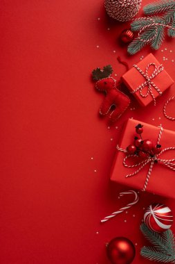 Dikey kompozisyonda, ipliğe sarılı hediye kutuları, kırmızı süsler, pelüş bir ren geyiği, şeker kamışı ve koyu kırmızı bir zemin üzerinde çam dalları bulunur.