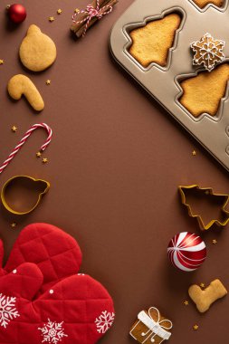 Dikey Noel pişirme sahnesinde bir tepsi ağaç şekilli kurabiye, şeker kamışı, eldiven, süsler ve sıcak kahverengi arka planda hediye aksanları yer alıyor.